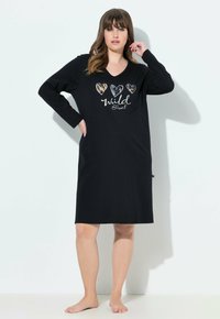 Frau, die barfuß steht und ein schwarzes langärmeliges knielanges Kleid trägt, auf dem drei gemusterte Herzen und der Text "wild @ heart" auf der Vorderseite zu sehen sind.