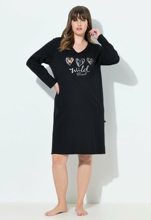 Donna in piedi a piedi nudi, che indossa un vestito nero a maniche lunghe fino al ginocchio con tre cuori decorati e il testo "wild @ heart" davanti.
