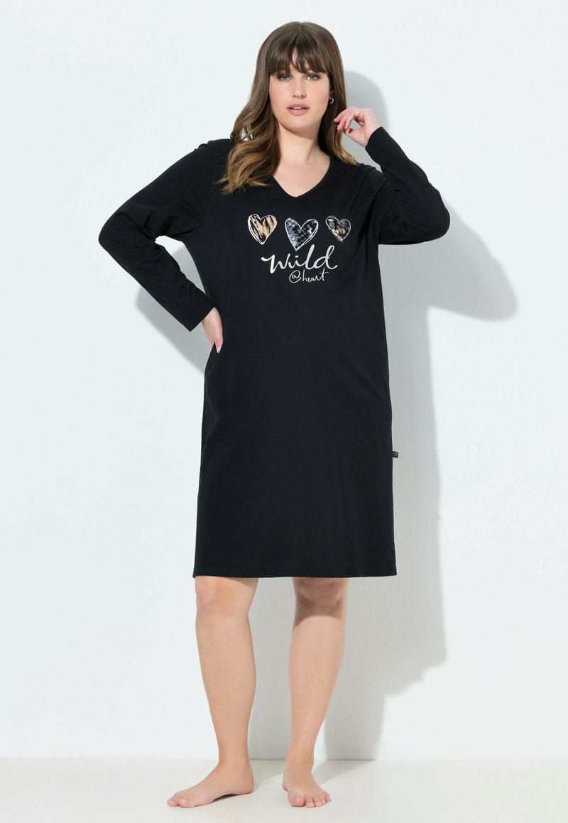 Frau, die barfuß steht und ein schwarzes langärmeliges knielanges Kleid trägt, auf dem drei gemusterte Herzen und der Text "wild @ heart" auf der Vorderseite zu sehen sind.