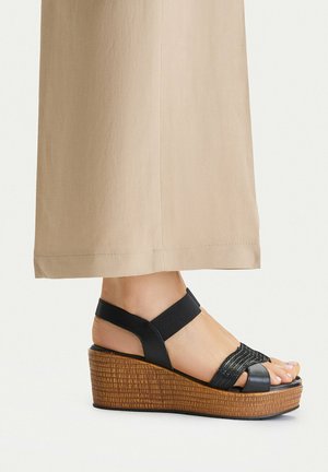 Pie con sandalia negra de cuña con tiras y plataforma marrón texturizada, combinada con pantalones anchos beige sobre un fondo claro y liso.