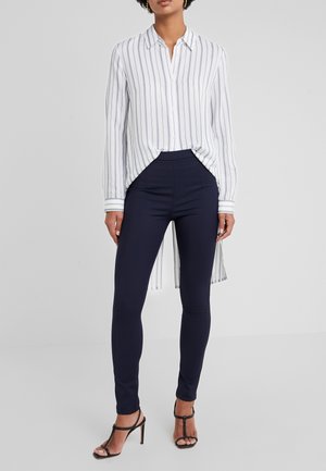 Kobieta w białej koszuli z długim rękawem z pionowymi paskami, granatowych dopasowanych spodniach i czarnych sandałach na wysokim obcasie stoi na tle szarego tła.