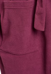 Bourgogne fleeceplagg med en texturerad yta, som har en stor ficka och detalj med knytning i midjan. Hållbart och mjukt material.