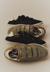Deux paires de baskets décontractées, une noire et une vert olive, toutes deux avec trois bandes velcro, disposées en chevauchement sur une surface beige.
