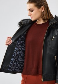 Giacca di pelle nera con rifiniture in pelliccia, accessori in oro e foderata con un motivo blu-grigio. Il modello indossa un maglione bordeaux sotto.