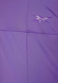 Lila träningslinne med en slät yta, subtila sömdetaljer och en reflekterande logotypaccent i det övre högra hörnet.