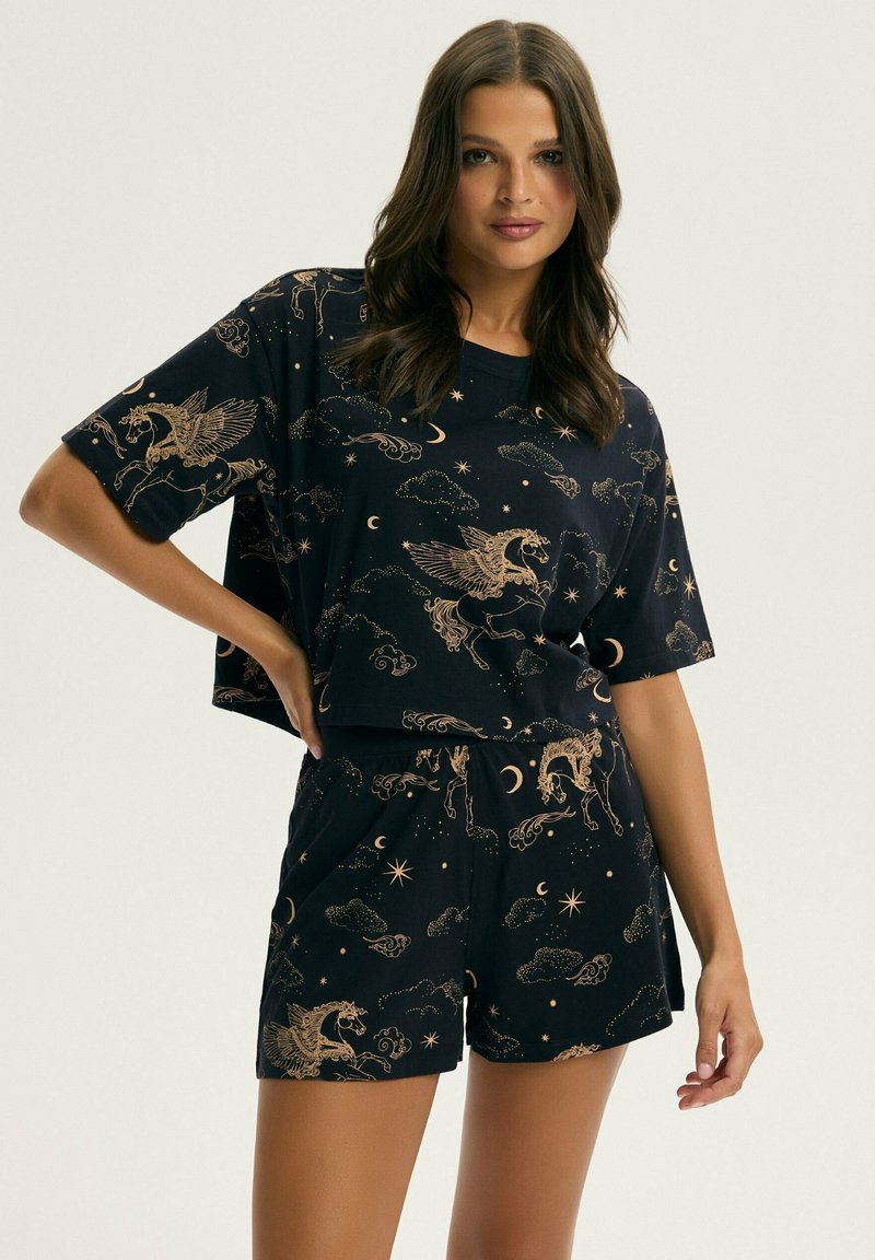 Kobieta ubrana w czarne krótkie spodenki pasujące do oversize'owej koszulki z motywem złotego Pegaza i gwiazd, stojąca na jednolitym tle.