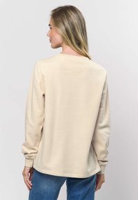 Gaiši bēša garroku džemperis ar apaļu kakla izgriezumu. Mīksta tekstūra, brīvs piegriezums un ribots aproču raksts. Kombinēts ar zilām džinsa biksēm.