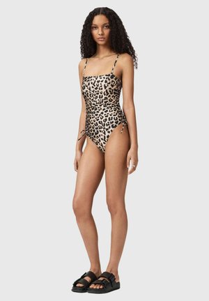 Kvinde med langt krøllet hår iført en leopardmønstret badedragt i ét stykke og sorte sandaler med spænde, stående foran en ensartet grå baggrund.