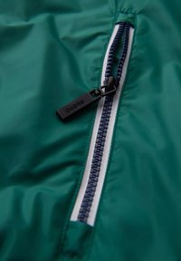 Grön nylonväv med en dragkedja i marinblått och svart dragflik, accentuerad med vit söm längs dragkedjan.