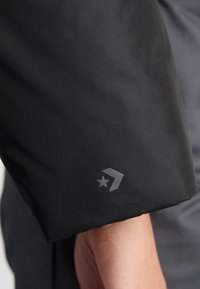 Čierna nepremokavá bunda, rukáv s hladkou textúrou. Obsahuje šedú hviezdnu logo a chevron symbol na manžete.