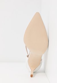 Une chaussure blanche à talon aiguilles posée sur le côté, montrant la semelle beige lisse et la petite pointe du talon sur un fond blanc.