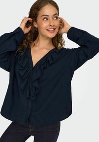 Jeune femme aux cheveux bouclés portant un blouse navy avec des volants, souriant et ajustant ses cheveux devant un fond uni.