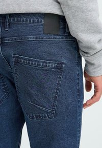 Blugi din denim albastru închis, cu o textură netedă, un petic din piele neagră la talie și buzunare mari, plasate în unghi, pe spate.