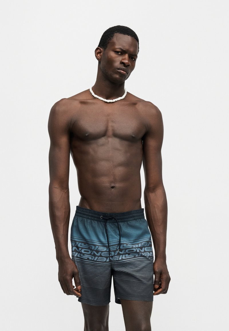 Άντρας χωρίς πουκάμισο που φοράει λευκό περιδέραιο με χάντρες και μπλε-μαύρα μαγιό με διαβάθμιση και κορδόνι, στέκεται μπροστά σε απλό ανοιχτό φόντο.