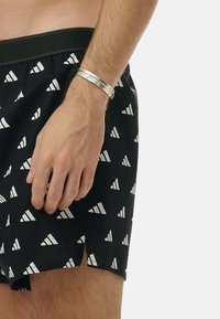 Svarta simshorts med vita triangulära mönster, med resårmidja. Ett silverarmband bärs på handleden.