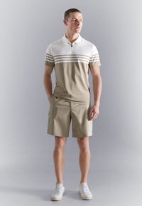 Hombre vestido con una camiseta tipo polo de manga corta beige y blanca con rayas, pantalones cortos cargo beige y zapatillas blancas, de pie sobre un fondo gris.
