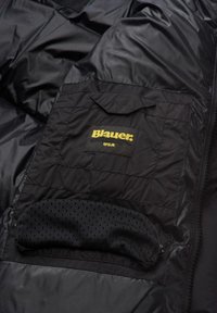 Giacca nera imbottita con una tasca frontale dotata di pattina e il logo "Blauer USA" in giallo. Include un dettaglio con tasca in rete.