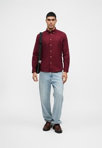 Muškarac stoji u bordo košulji s dugim rukavima, svijetloplavim trapericama, smešnim cipelama i nosi crnu torbu preko ramena.