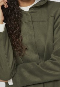 Olivgrön jacka med mjuk textur, huva och en krämfärgad stickad krage. Har synliga sömmar och en öppen frontdesign.