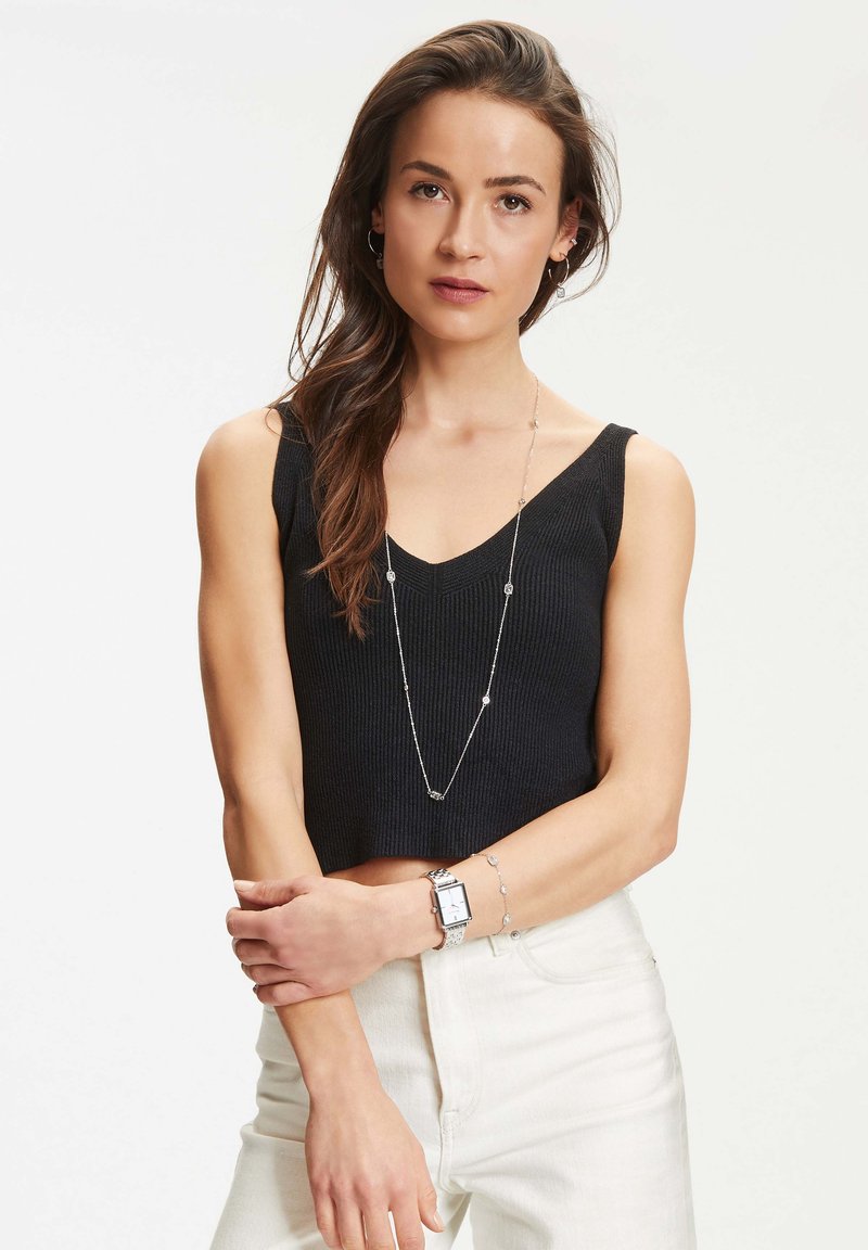 Schwarzes geripptes Crop-Top mit V-Ausschnitt, kombiniert mit weißen hochgeschnittenen Jeans. Zubehör umfasst eine lange silberne Halskette und eine silberne Uhr.