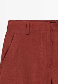 Pantalon en lin couleur rouille avec un design à plat devant, des poches latérales et une finition texturée. La ceinture est équipée d'une petite boucle.