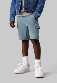 Helle blaue Cargo-Shorts aus weichem Stoff mit Seitentaschen. Kombiniert mit weißen Sneakers und gerippten weißen Socken sowie einer Jeansjacke.