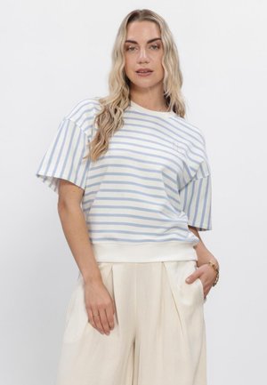 Vrouw met lang blond haar die een lichtblauw-wit gestreept shirt met korte mouwen en crèmekleurige, hooggetailleerde wijde broek draagt.