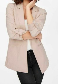 Ljusbeige kavaj med skräddarsydd passform, flikiga slag, upprullade ärmar och två sidofickor, bärs över en vit topp och svarta byxor.