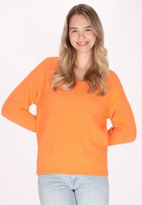 Pulover tricotat portocalie cu decolteu în V, mâneci lungi și o croială relaxată. Purtat cu blugi de culoare bleu deschis, expus pe un fundal neutru.