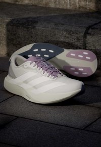 Paire de chaussures de course blanches avec des lacets violets clairs et des accents sur la semelle, posées sur un pavé en pierre près d'escaliers sombres.