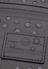 Tessuto grigio testurizzato con gocce d'acqua, presenta un'etichetta in gomma nera con il logo in rilievo "JOHNNY URBAN" e elementi decorativi.