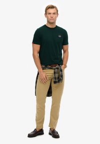 Camiseta de algodón de color verde oscuro con un pequeño logo, pantalones beiges y una camisa de franela a cuadros atada en la cintura. Zapatos de cuero marrón.