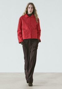 Red cropped jacket with two front pockets, button closure, and a pointed collar, paired with loose dark brown trousers and brown boots.