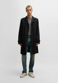 Schwarzer langer Mantel, graues T-Shirt, blaue abgenutzte Jeans und beige Sneaker. Das Modell steht mit den Händen an den Seiten vor einem hellen Hintergrund.