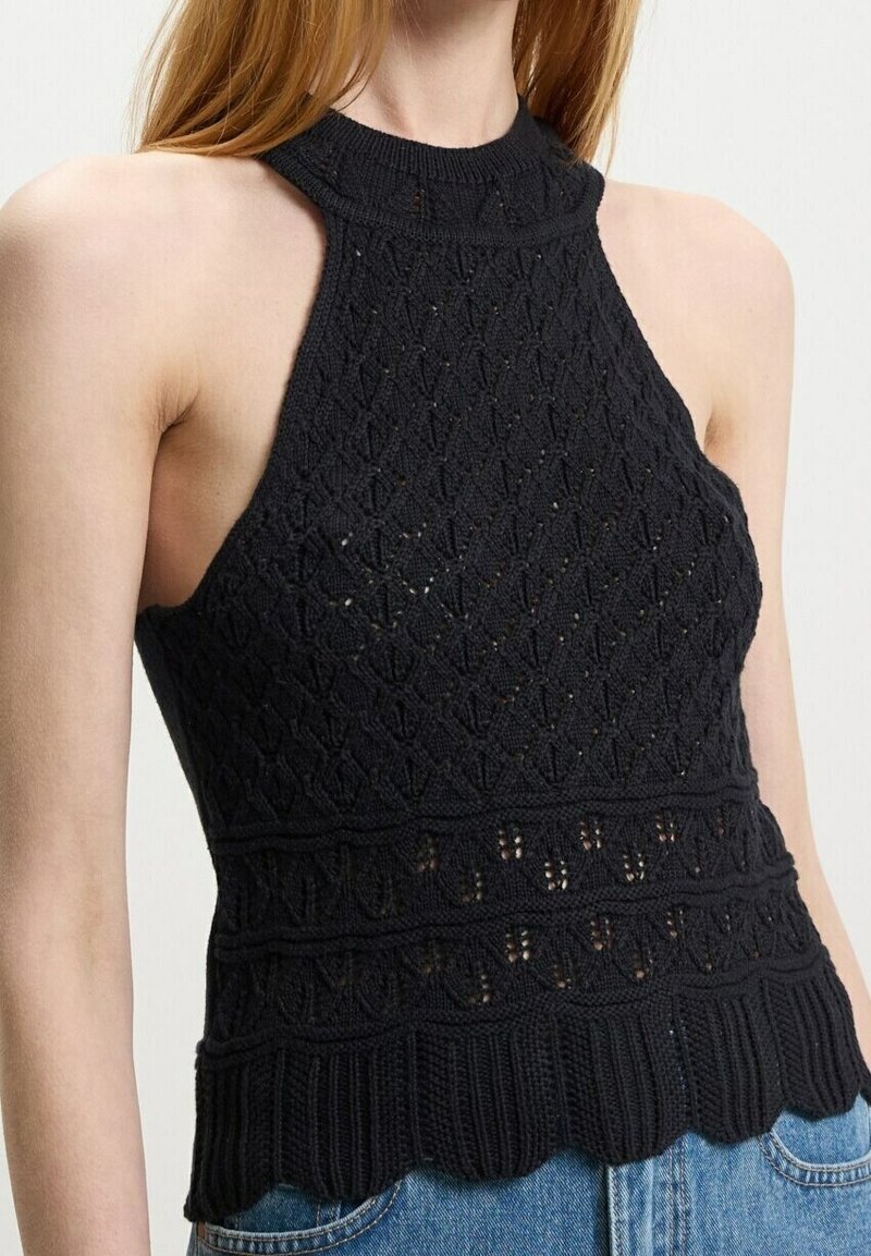Femme portant un top dos nu noir en maille avec une texture à motifs et un ourlet festonné, associé à un jean bleu.
