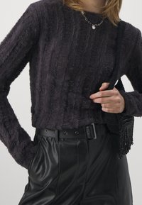 Person, die einen dunklen, flauschigen Pullover, schwarze Lederhosen mit Gürtel trägt und eine schwarze gestrickte Schultertasche mit Fransen hält.