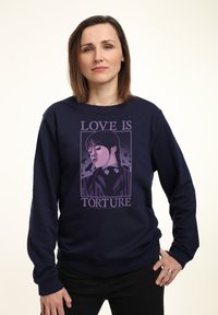 Donna con capelli castani di lunghezza spalle, che indossa una felpa blu marina con una grafica viola e il testo "L'AMORE È TORTURA", con la mano sulla hip.