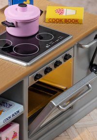 Miniaturowy zestaw kuchenny z drewnianym blatem, czarną płytą grzejną, różowym garnkiem, srebrnym piekarnikiem i kolorowymi książkami kucharskimi. Widoczne szczegółowe elementy i tekstury.