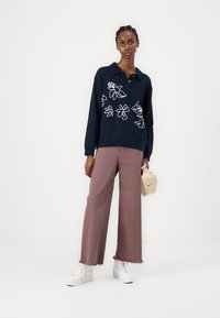 Mornarsko pulover s belim cvetličnim vezenjem in ovratnikom, v kombinaciji s svetlo roza širokimi hlačami in belimi športnimi copati, drži majhno kremasto torbico.