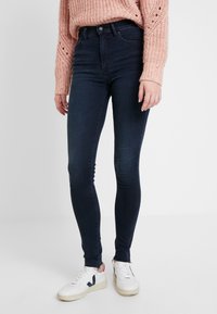 Jeans skinny de denim azul oscuro con una cintura alta, que presentan una textura suave y un ajuste entallado, combinados con zapatillas blancas.