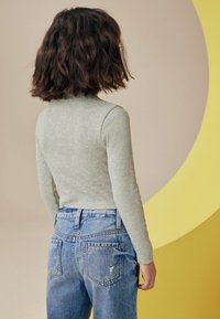 Grå langærmet ribbet top parret med højtaljede lyseblå jeans. Baggrunden har en stor gul cirkulær form.