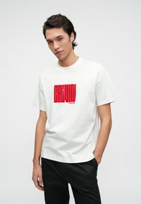 White cotton t-shirt featuring a bold red graphic print with "CALVIN KLEIN" text. Short sleeves and a round neckline. Worn with black trousers.