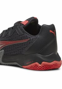 Zapatilla deportiva negra con parte superior de malla texturizada, acentos sintéticos y una suela roja con un patrón en zigzag. Marca destacada en el talón.