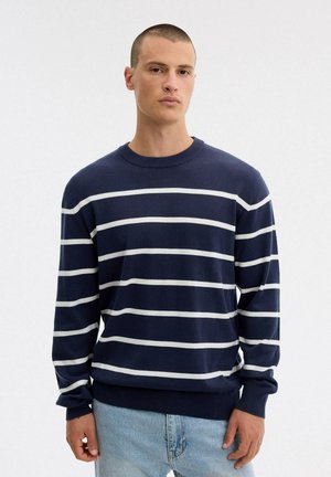Un hombre posa con un jersey azul marino de rayas horizontales blancas y unos vaqueros azul claro sobre un fondo blanco liso.