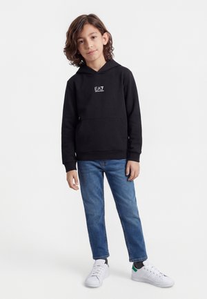 Enfant debout portant un sweat à capuche noir EA7, un jean bleu et des baskets blanches sur un fond blanc uni.