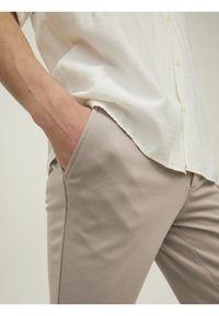 Beige nauwsluitende broek van een gladde stof, met zijzakken. Over de taille hangt een lichtgekleurde, doorgestikte blouse.