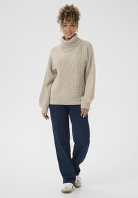Beiger Strickpullover mit hohem Rollkragen, ausgestattet mit einem Zopfmuster. Kombiniert mit dunkelblauen Jeans und weißen Sneakers.