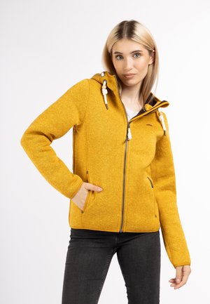 Jachetă din polar galbenă, cu fermoar și glugă, cu țesătură texturată, accente cu șnur și buzunare laterale. Stilizată cu blugi negri.