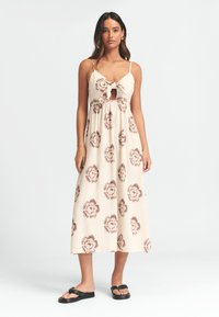 Maxi šaty s květinovým vzorem v hnědé barvě na krémovém pozadí. Mají zavazování na přední části a výřez. Nosí se s plochými černými sandály.