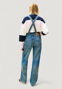 Jeansowy kombinezon w niebieskim odcieniu z regulowanymi paskami na krzyż z tyłu oraz dwoma kieszeniami z tyłu. Do tego wielokolorowa, paskowana bluza.